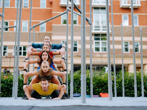 A group of six performers in bright clothes balance on top of one another, in between metal scaffolding.