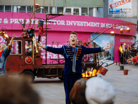 A performer in a blue and red band uniform has his arms outstretched, with a truck and a bin of fire in the background.