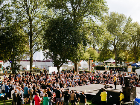 An audience gather around a stage, with trees in the background.