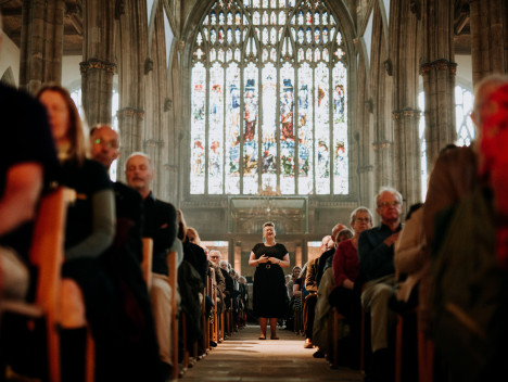A singing woman walks through the centre aisle of a church, with the audience seated on either side. Large stained glass windows are in the background.