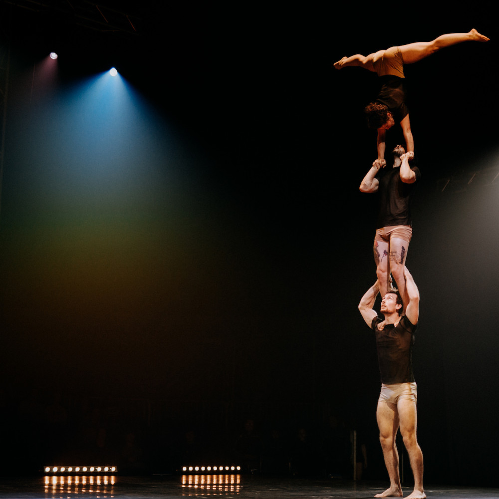 Three performers are balanced on top of one another: the performer at the top is upside down, doing the splits. Another performer looks on.